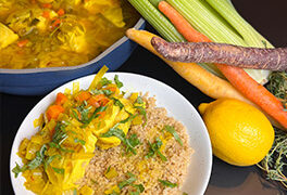 Poached chicken in a saffron, leek and parsley broth is served over farro on a white plate with a blue pot full of the entree in the background.