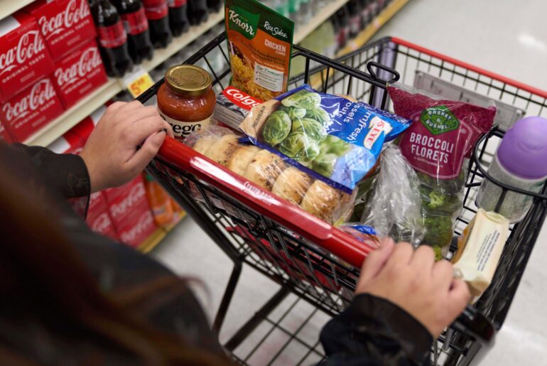 A person pushes a shopping cart containing groceries such as rolls, pasta sauce, spinach, broccoli, toothpaste, and instant soup in a supermarket aisle.