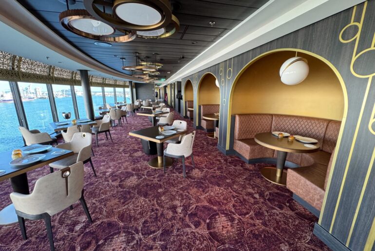 Elegant restaurant interior with arched booths, round tables, and plush seating. Large windows offer a view of water and buildings. The decor features gold accents and modern lighting that bloom with sophistication.