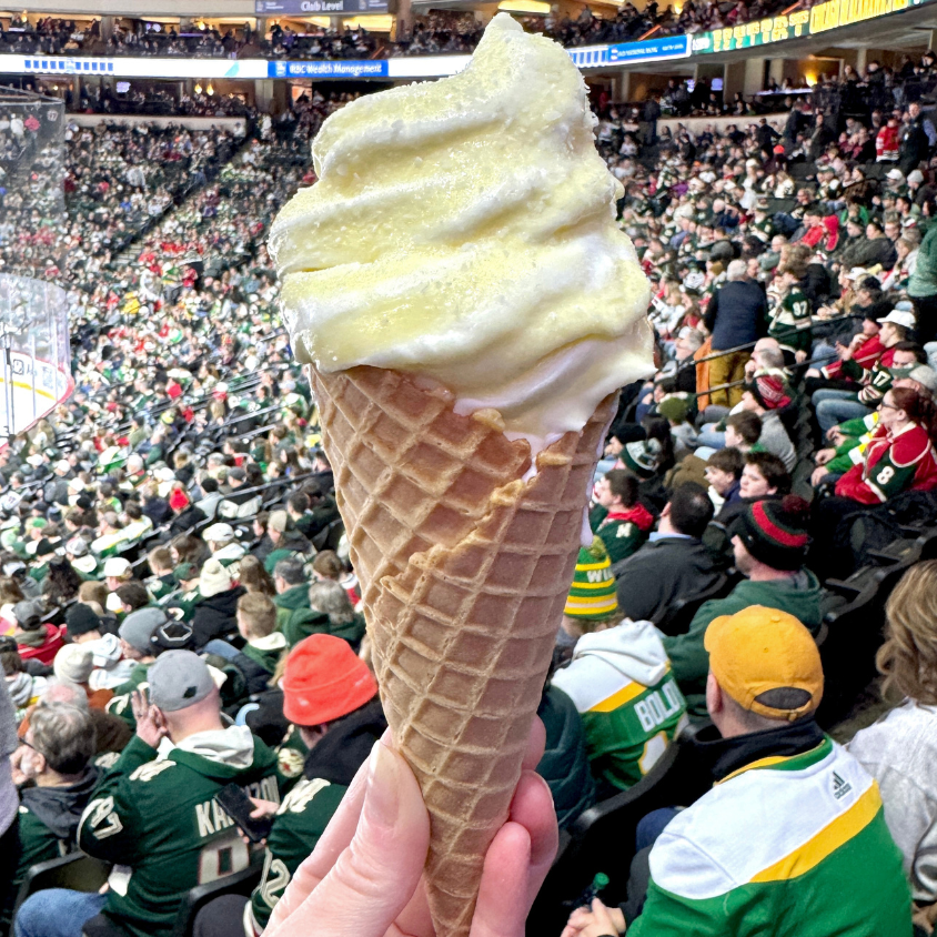 A hand holding a large ice cream cone in a crowded sports arena. A hand holding a large ice cream cone in a crowded sports arena.