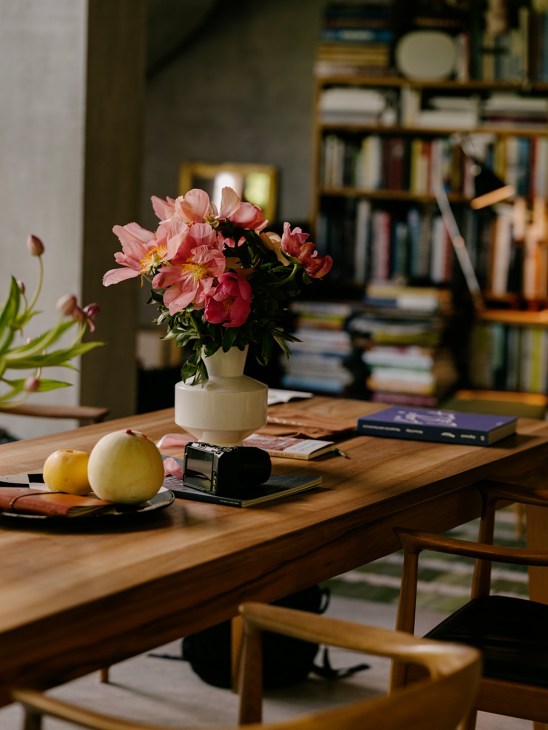 Wooden desk with pink peonies in white vase, apples, books, and homeware against blurred bookshelves.