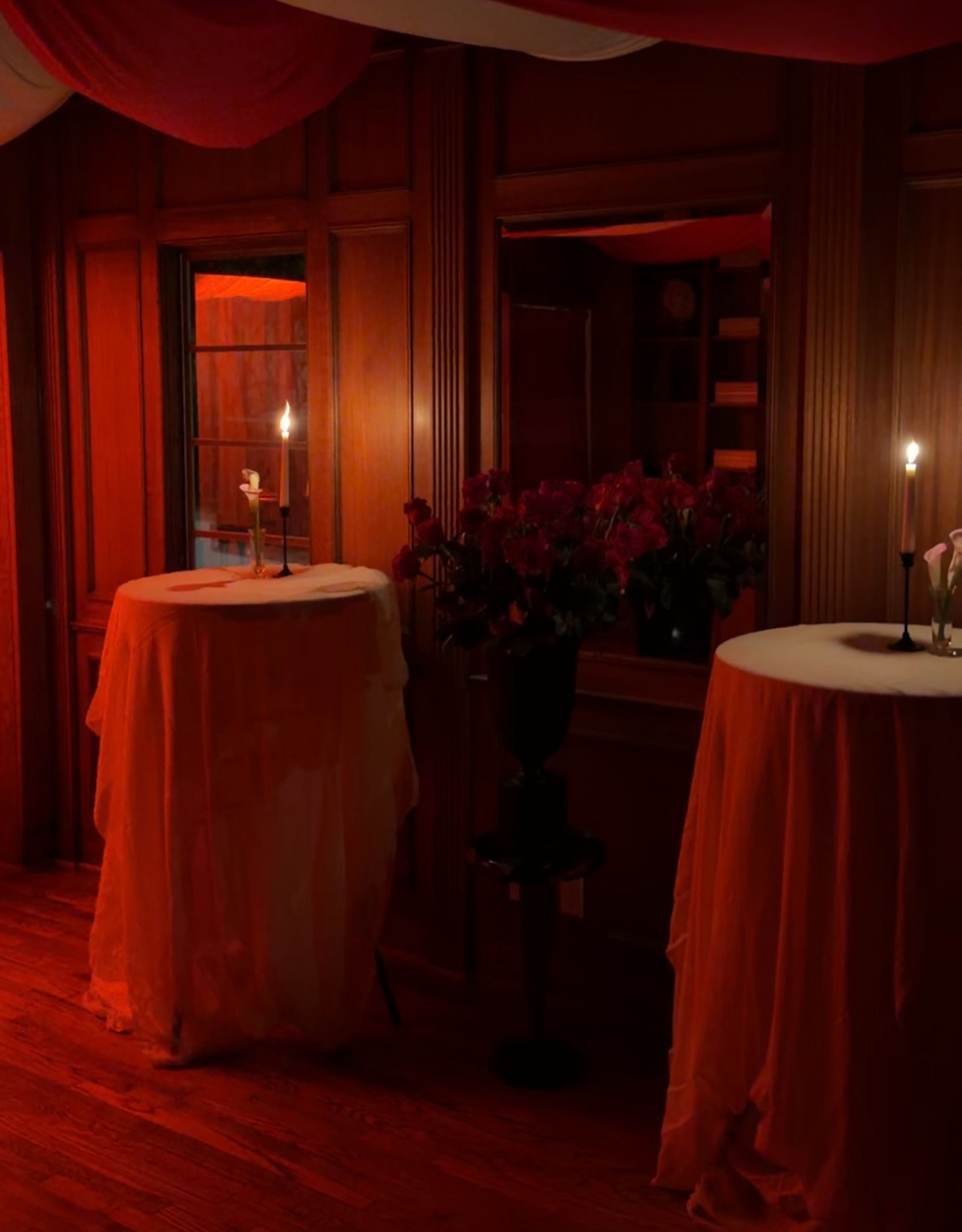 A bar lounge decorated for halloween with everything lit up red and two bar tables covered in tablecloths with lit taper candles on top